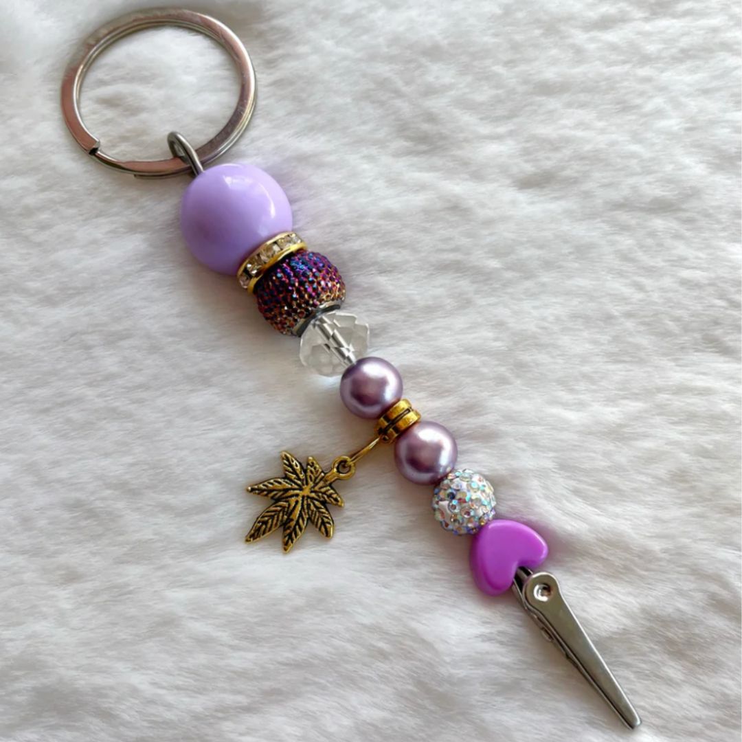 Purple and gold beaded roach clip with rhinestones and a gold cannabis leaf charm, handmade by Trap Mami LA.