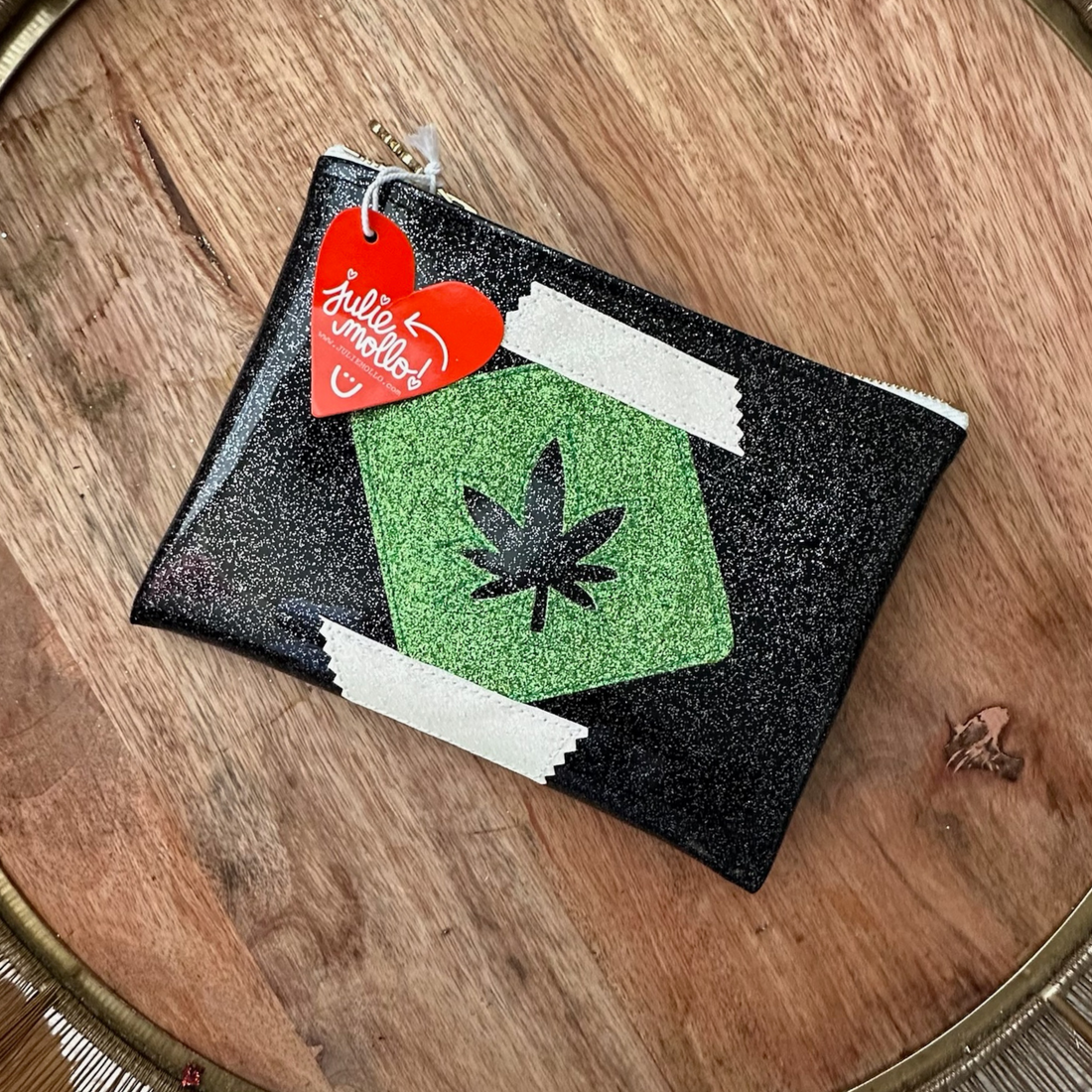 A black glittery pouch with a green pot leaf cut out of the center. The pouch has a zipper closure and a red heart-shaped tag that reads &quot;Julie Mollo&quot; attached to the zipper pull. It rests on a wooden surface.