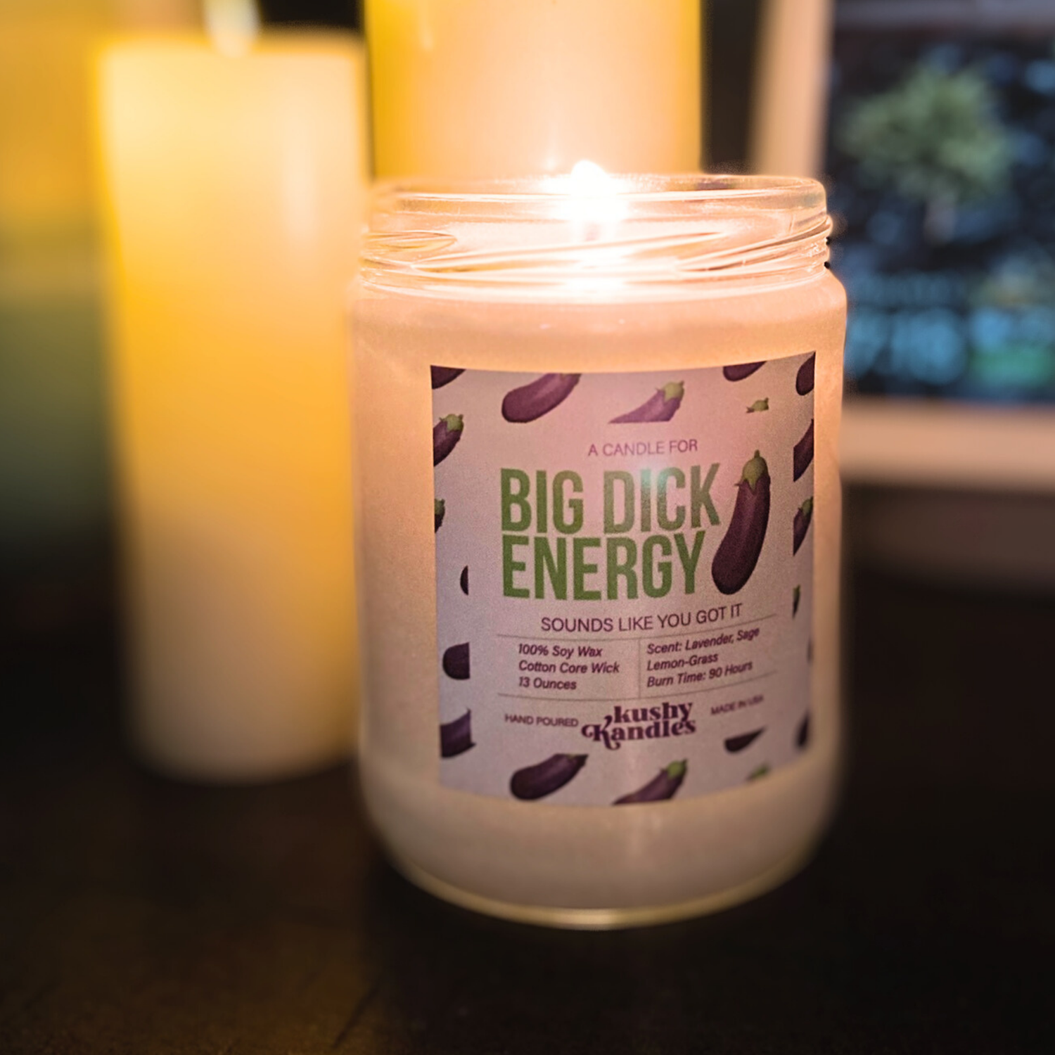 A glowing candle labeled &quot;Big Dick Energy&quot; surrounded by soft ambient lighting. The candle sits in a cozy setting, showing its lavender, lemongrass, and sage scent on the label with an eggplant emoji design.