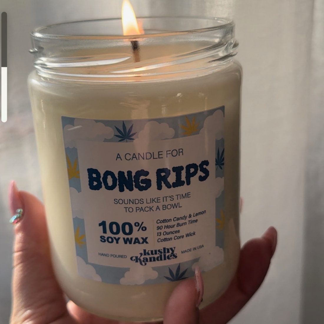 A hand holding a lit &quot;Bong Rips&quot; soy wax candle with cotton candy and lemon scent, bringing playful and relaxed vibes in a soft-lit room.