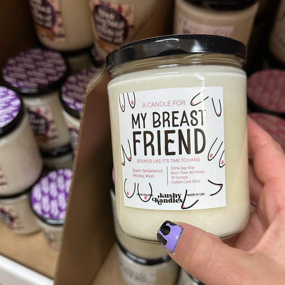 A hand holding the &quot;My Breast Friend&quot; soy wax candle with sandalwood and amber scent, displayed against a backdrop of other candle products, evoking friendship and warmth.