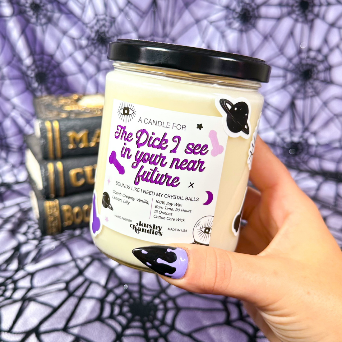 A hand holding the Future Dick Vanilla &amp; Lemon Soy Wax Candle with creamy vanilla and lemon scent, set against a Halloween-themed backdrop with spider webs and books.