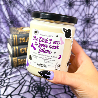 A hand holding the Future Dick Vanilla &amp; Lemon Soy Wax Candle with creamy vanilla and lemon scent, set against a Halloween-themed backdrop with spider webs and books.