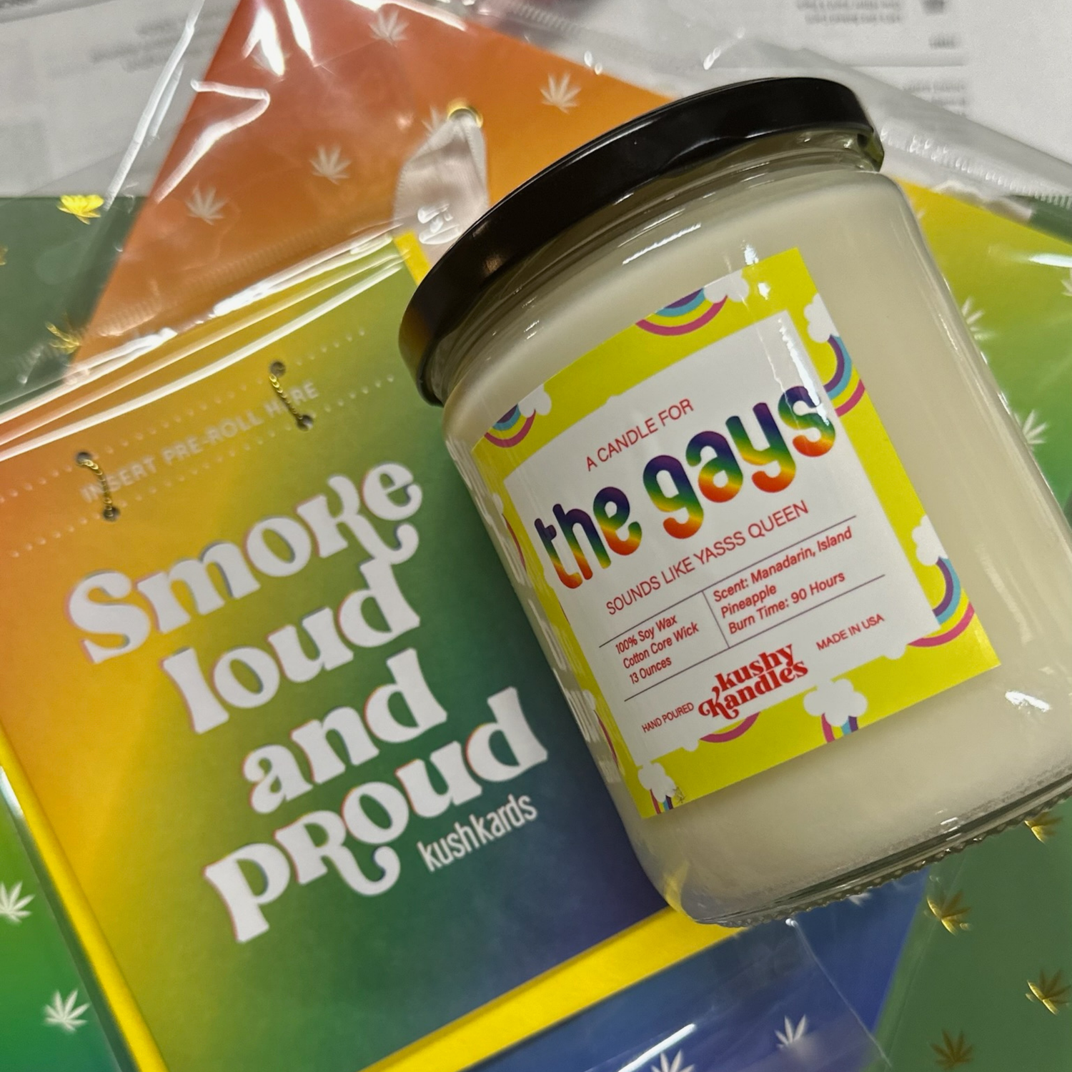 A &quot;The Gays&quot; soy wax candle with a mandarin and pineapple scent displayed against a colorful background with the words &quot;Smoke Loud and Proud&quot; in the backdrop.