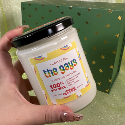 A hand holding the &quot;The Gays&quot; soy wax candle with a mandarin and pineapple scent in front of a green box decorated with gold cannabis leaves, evoking pride and celebration.