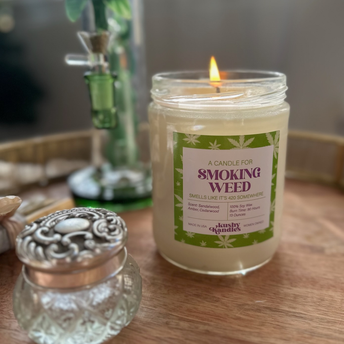 A lit &quot;Smoking Weed&quot; soy wax candle in a cozy indoor setting, featuring a sandalwood and amber scent, next to elegant home decor and a green bong, evoking a relaxed, serene atmosphere.