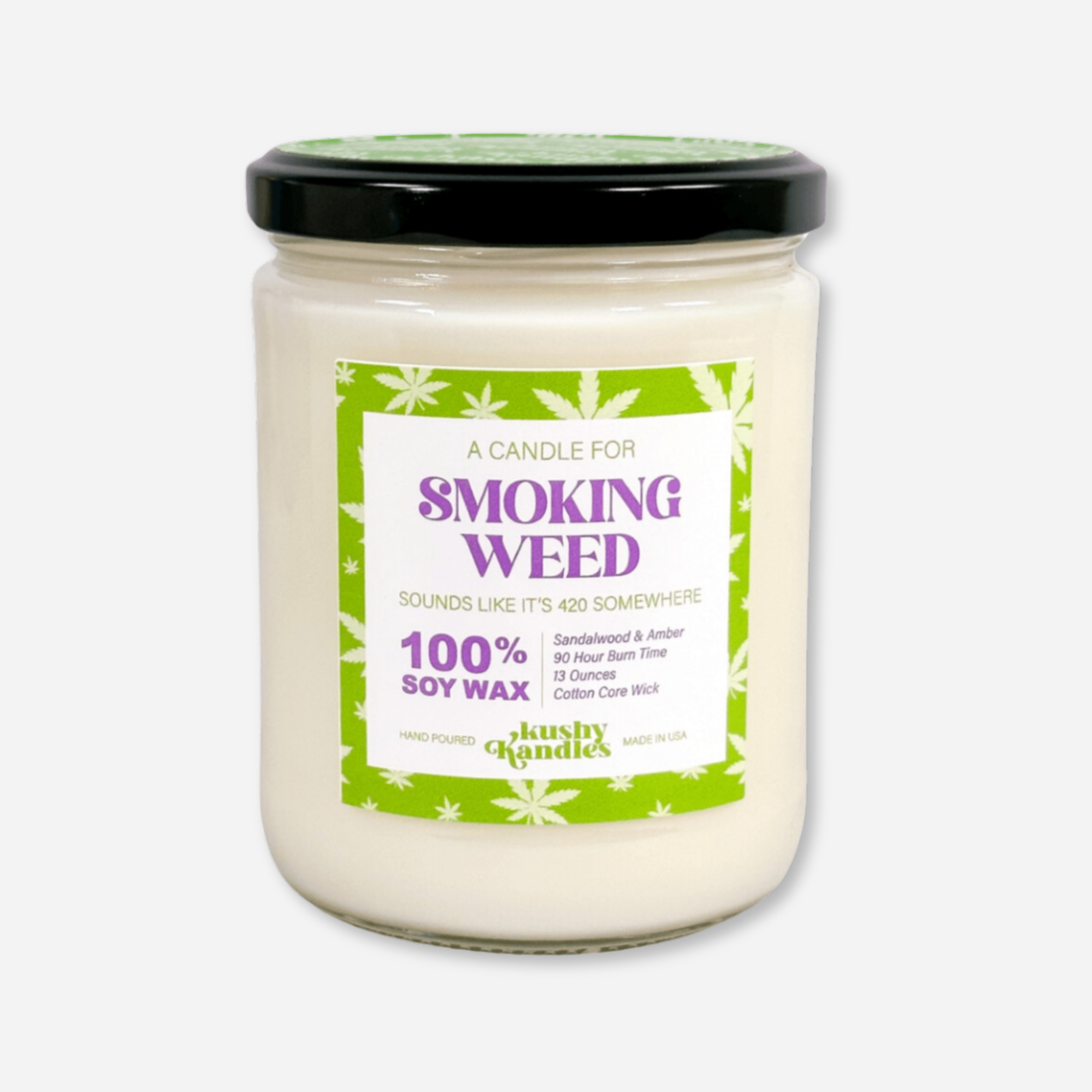 A clean product shot of the &quot;Smoking Weed&quot; soy wax candle in a glass jar with a black lid. The label highlights sandalwood and amber as the primary scents, with a 90-hour burn time and a 100% soy wax formula, surrounded by a green cannabis leaf design. ​