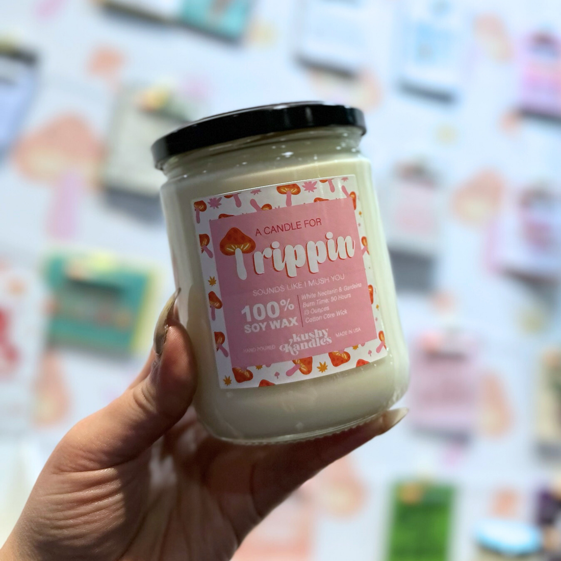 A hand holding the &quot;Trippin&quot; soy wax candle with white nectarine and gardenia scent against a blurred backdrop of colorful, playful products.